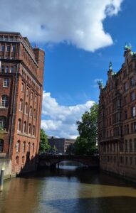 Foto des Nikolaifleets in Hamburg. An den Seiten des Fleets ragen die typischen Häuser der Speicherstadt mit ihrem roten Klinker hoch. Der Himmel ist blau und wird von ein paar Wolken bedeckt.