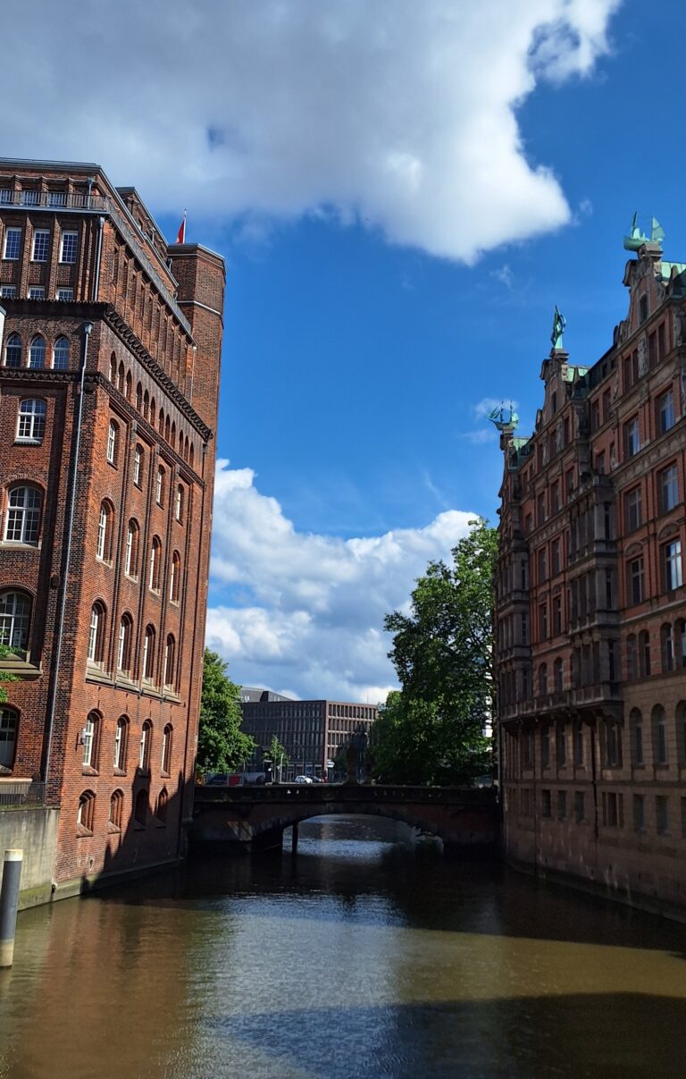Foto des Nikolaifleets in Hamburg. An den Seiten des Fleets ragen die typischen Häuser der Speicherstadt mit ihrem roten Klinker hoch. Der Himmel ist blau und wird von ein paar Wolken bedeckt.