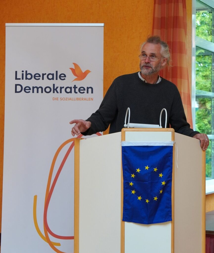 Der Vorsitzende des Landesverbands Niedersachsen, Steve Bourne, steht am Rednerpult und spricht zu den anwesenden Parteimitgliedern. Das weiße Rednerpult wurde mit einer EU-Flagge dekoriert, im Hintergrund steht ein Werbeaufsteller mit dem Logo der Liberalen Demokraten. 